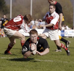 Rugby-Spieler fällt mit Ball über die Linie auf die Kamera zu, während hinter ihm mehrere Spieler angerannt kommen