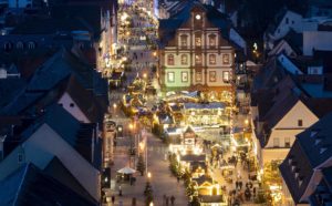 Ausschnitt aus einer Aufnahme aus einem der Domtürme auf die Maximilianstraße mit beleuchteten Weihnachtsmarktständen bei Dämmerung