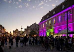 Viele Menschen auf der Maximilianstraße, rechts das lila beleuchtete Sophie-de-la-Roche-Haus, im Hintergrund Alte Münze und Altpörtel