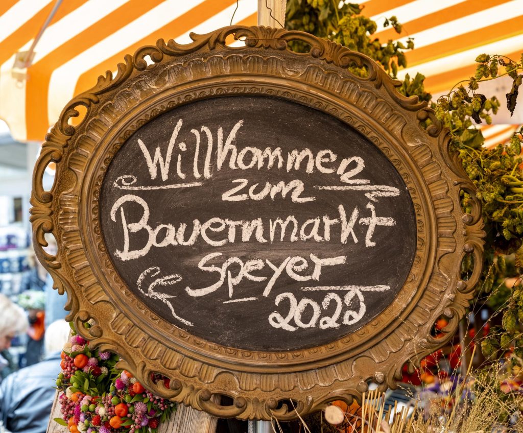 Urigens Schild mit der in Kreide gemalten Aufschrift "Willkommen zum Bauernmarkt Speyer 2022"