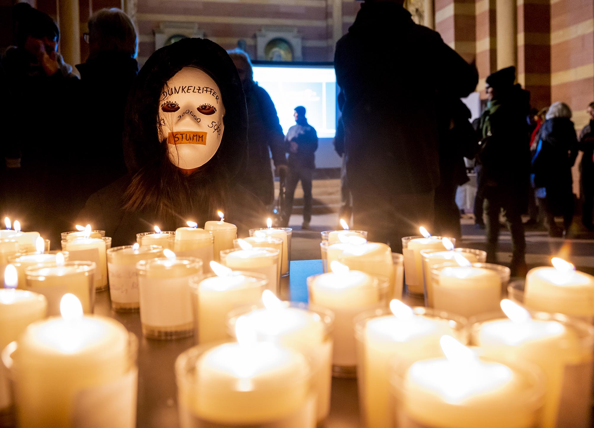 Viele brennende Kerzen und dahinter Gestalt mit weißer Maske, zugeklebtem Mund und Aufschrift Dunkelziffer im Kapuzenumhang