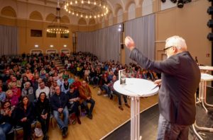 Autor Uwe Ittensohn prostet den zahlreichen Zuhörern im Alten Stadtsaal von der Bühne aus zu