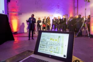 Laptop mit Präsentation im Vordergrund, im Hintergrund Menschen und Vortragender vor Leinwand