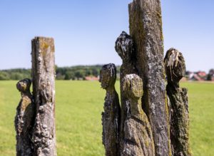 Verwittertes mit Flechten bewachsenes Holzkunstwerk in Form von stilisierten Menschenfiguren am Weg
