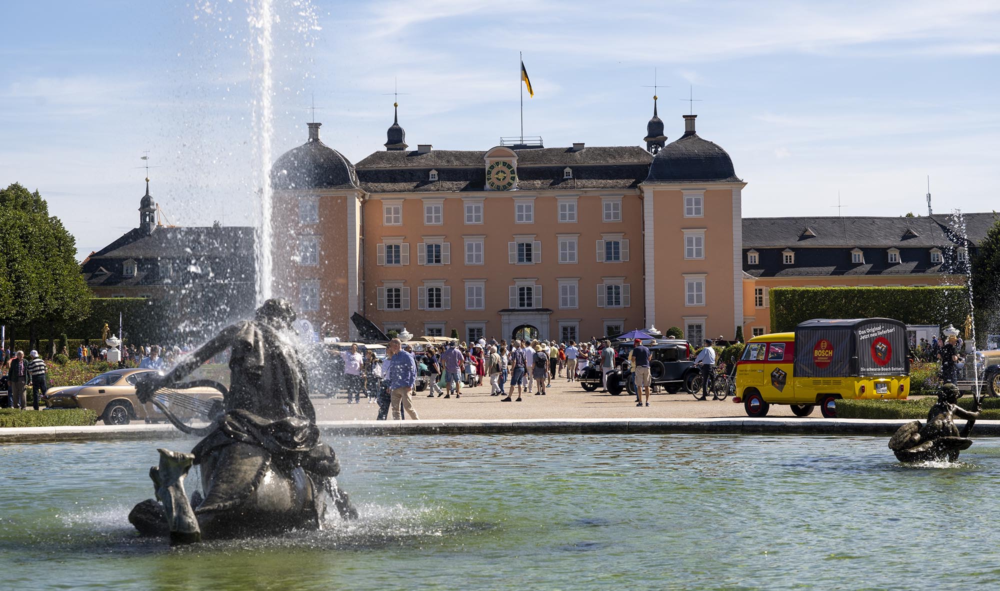 Schwetzinger Schloss durch eines der Wasserspiele im Schlossgarten fotografiert. Viele Menschen auf den Wegen, die sich die ausgestellten Autos anschauen.