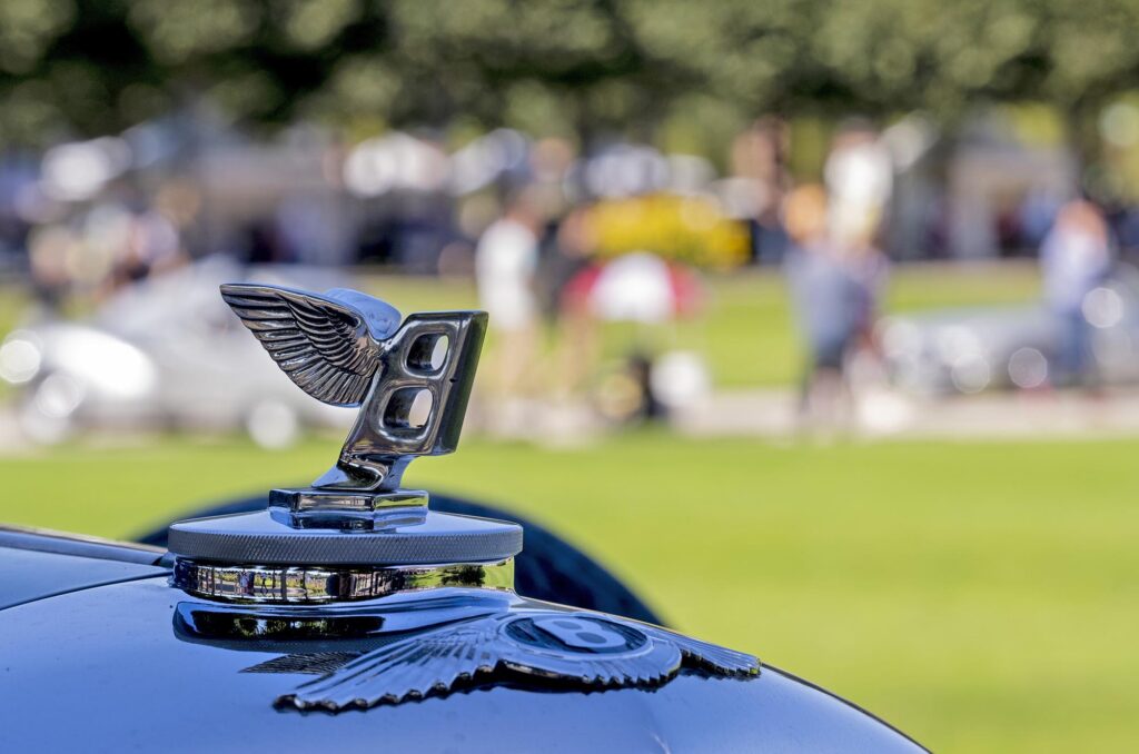 Geflügelte Bentley-Kühlerfigur in Nahaufnahme, dahinter unscharf im Hintergrund Grünanlage mit vielen bunt gekleideten Menschen