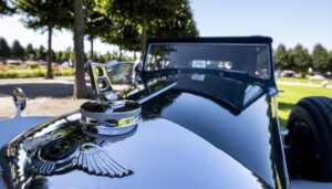 Nahaufnahme des geflügelten Bentleyzeichens auf einer spiegelblank geputzten schwarzen Motorhaube, in der sich der blaue Himmel spiegelt