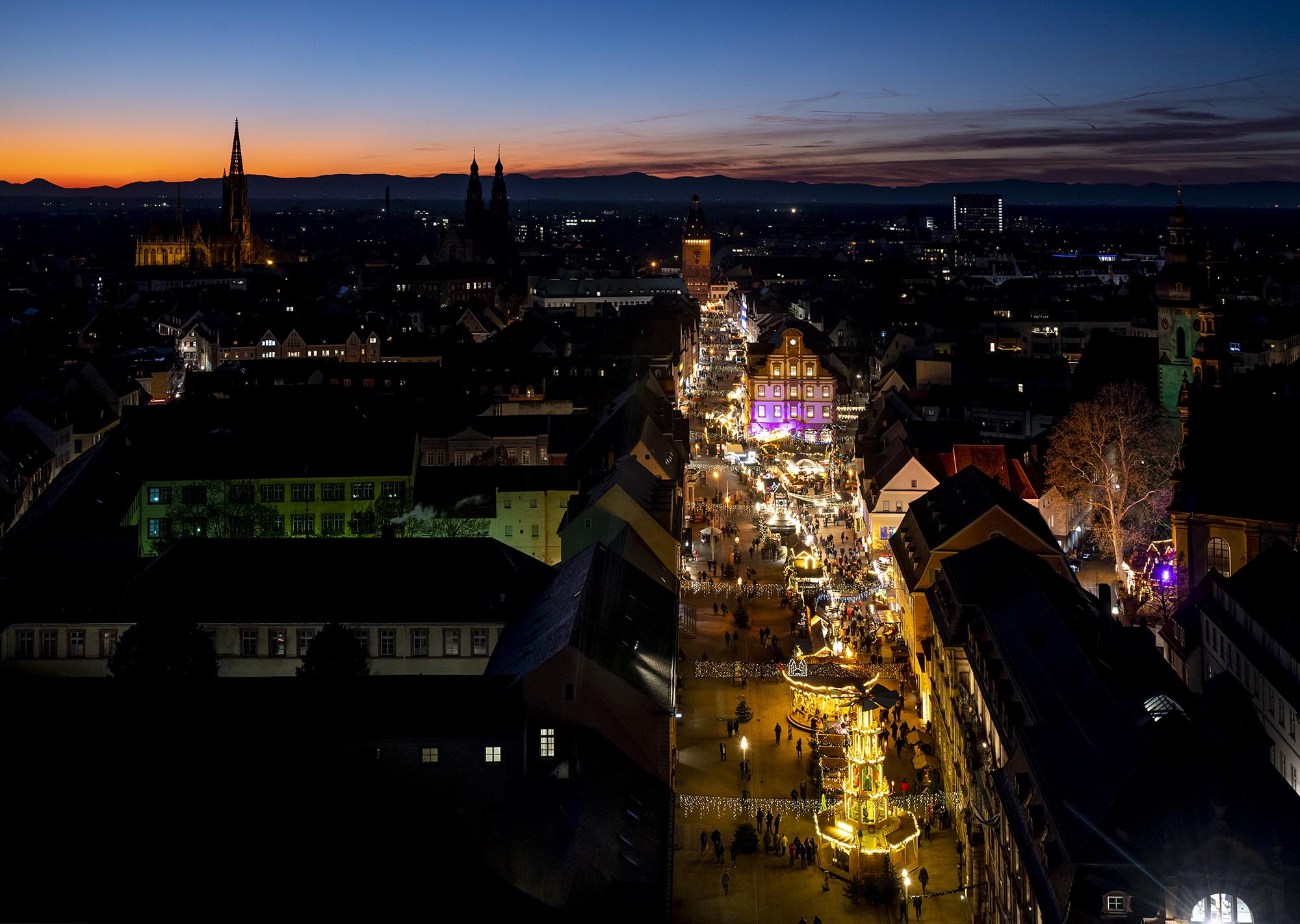 Blick vom Dom über die in der Dunkelheit bunt beleuchtete Maximilianstraße mit abendlichrm Neujahrsmarkt, Sonnenuntergang am Horizont