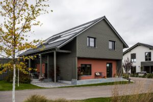 Rot gestrichenes Haus mit Metallsockel und schwarzer Lamellenfassade, unten teilweise, oben komplett, Solarpanele auf dem Dach.