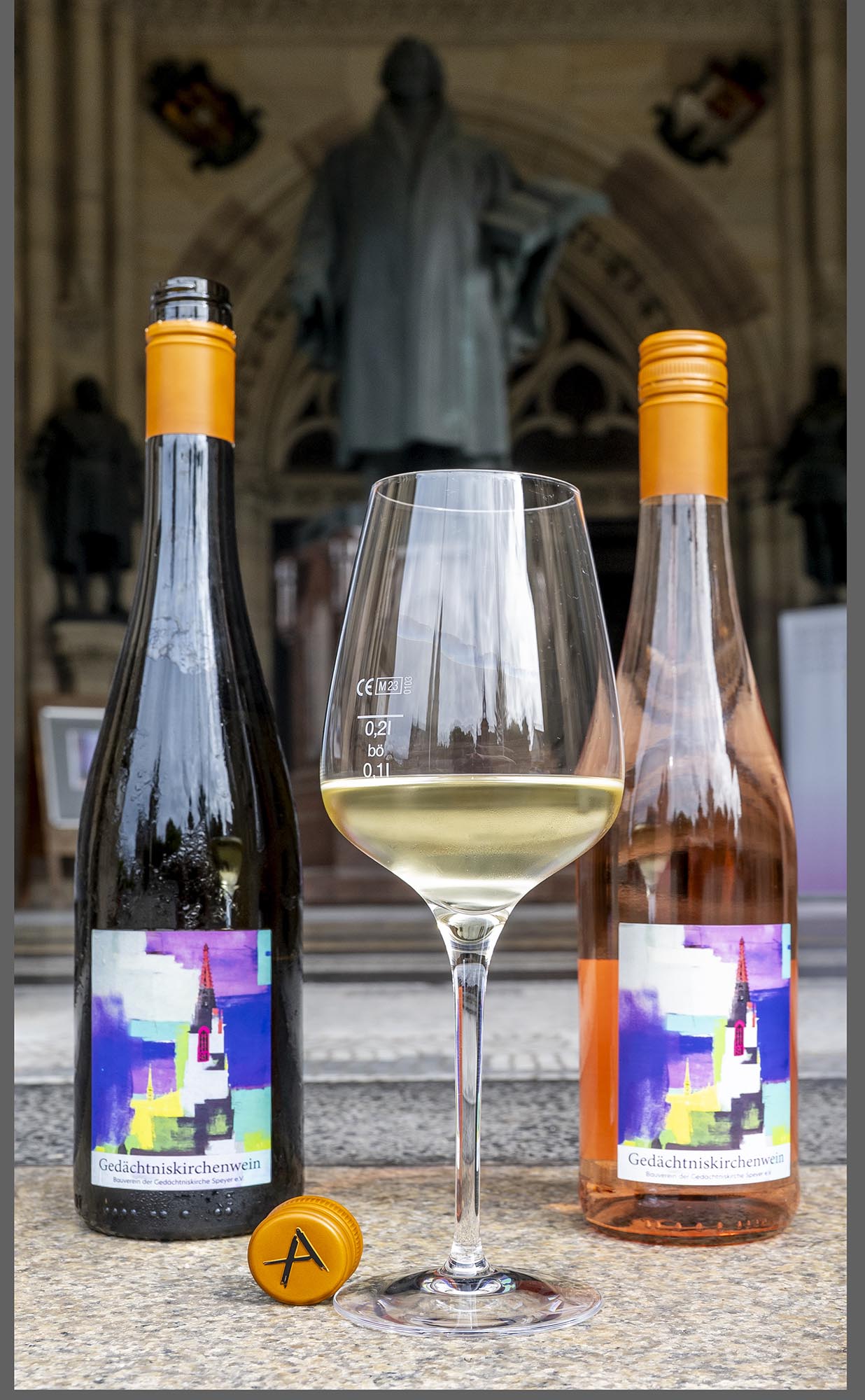 Zwei der Weinflaschen mit Künstleretikett und einem Glas Wein auf den Stufen der Gedächtniskirche. Im Hintergrund die Statue von Martin Luther