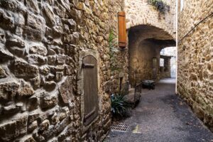 Blick durch eine verschlungene, mit breiten Torbögen teilweise überdachte Gasse mit Holztüren und vereinzelten Pflanzen