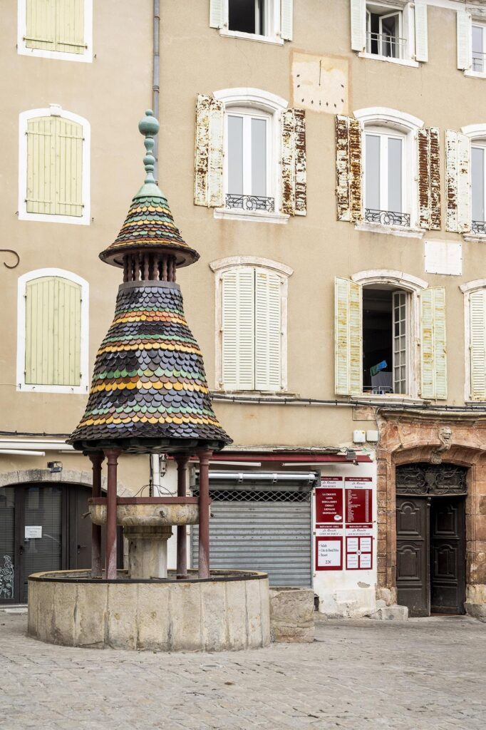 Mit bunten glänzenden Schindeln überdachter Sansteinbrunnen vor einem alten Haus mit wettergegerbten Fensterläden