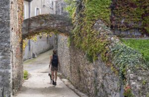 Mit wildem Wein überwachsener Torbogen in einer Gasse zwischen Natursteinhäusern. Eine Frau mit Hund läuft die Straße hoch.