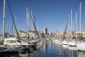 Im Hafen von Marseille liegende weiße Boote, die sich im blauen Wasser spiegeln. Im Hintergrund schicke Hafengebäude.