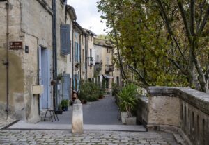 Malerische Gasse mit beigefarbenen Sandsteinhäusern mit blauen Fensterläden links und Bäumen rechts