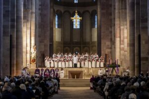 Blick durch das abgedunkelte Mittelschiff des voll besetzten Speyerer Doms zum beleuchteten Altarraum mit den Kirchenmännern und den Chorsängern