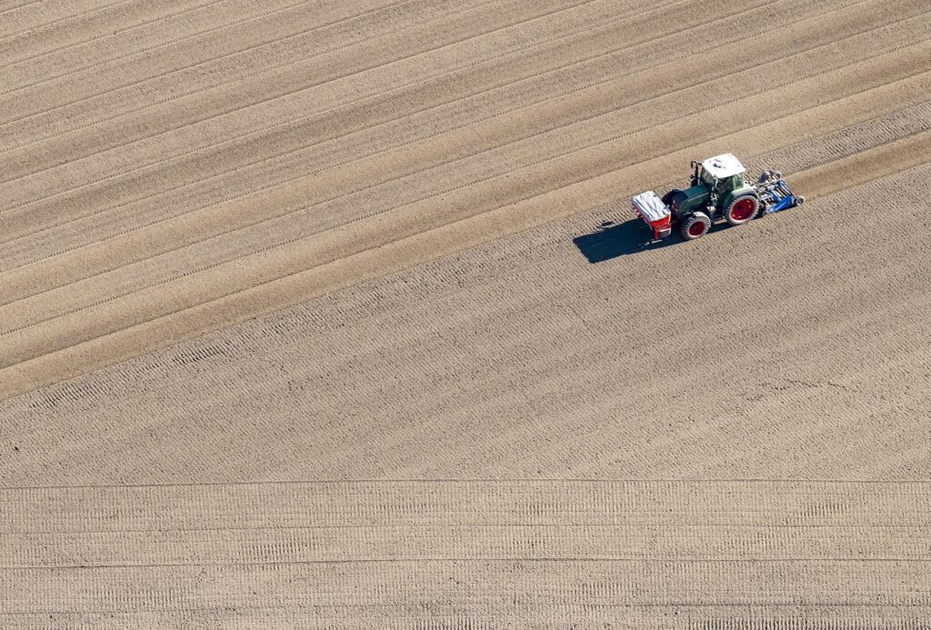Luftbild von einem frisch gepflügten, noch kahlem Feld, auf dem ein Traktor seine Linien zieht und die Ausaat macht.