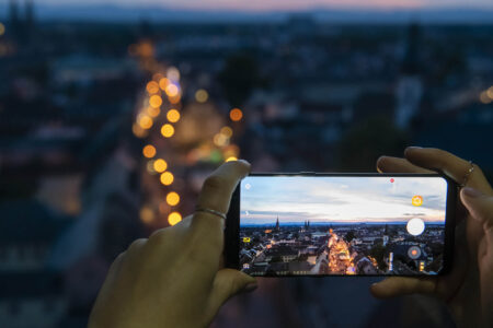 Frauenhände halten Smartphone, das die beleuchtete Maximilianstraße fotografiert, die man im Hintergrund verschwommen in groß sieht